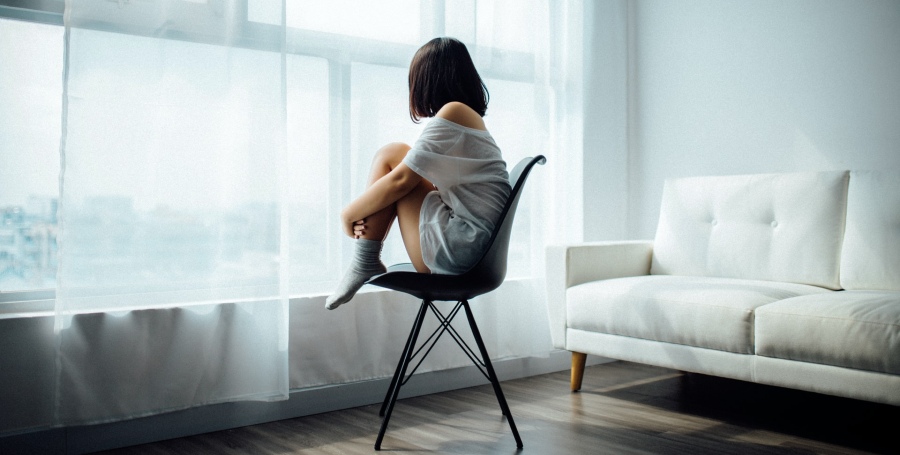 Depressed girl sits with legs up on a chair while sadly looking out the window